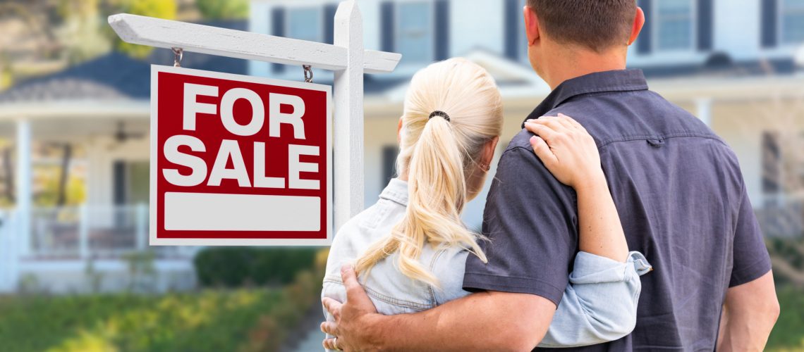 Young Adult Couple Facing Front of For Sale Real Estate Sign and House.